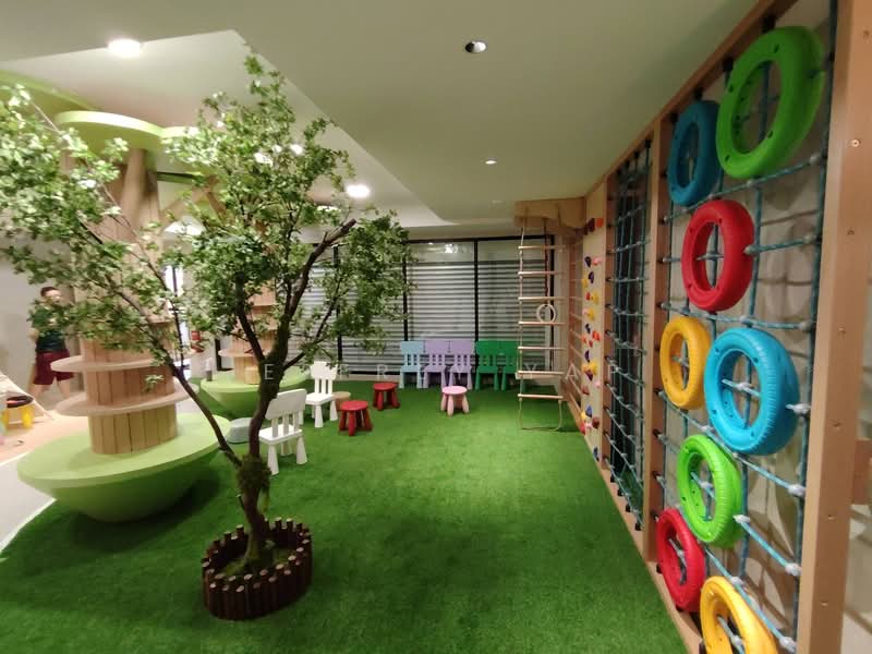 Room features: artificial grass flooring, colorful climbing wall with tires, wooden play structures, green tree decoration, small white chairs, small purple chairs, small pink chairs, small blue chairs, safety mats, indoor playground equipment, sliding door, overhead lighting; style: modern.