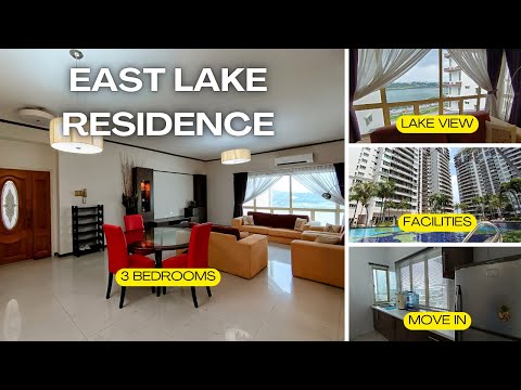 Living features: lake view through large windows, cream tiled flooring, recessed ceiling lights, 5-blade ceiling fan, L-shape beige fabric sofa, glass dining table with black legs, 4 red upholstered dining chairs, wall-mounted air conditioning unit, decorative wall paneling, curtains with sheer and blackout layers; style: modern; view: lake.