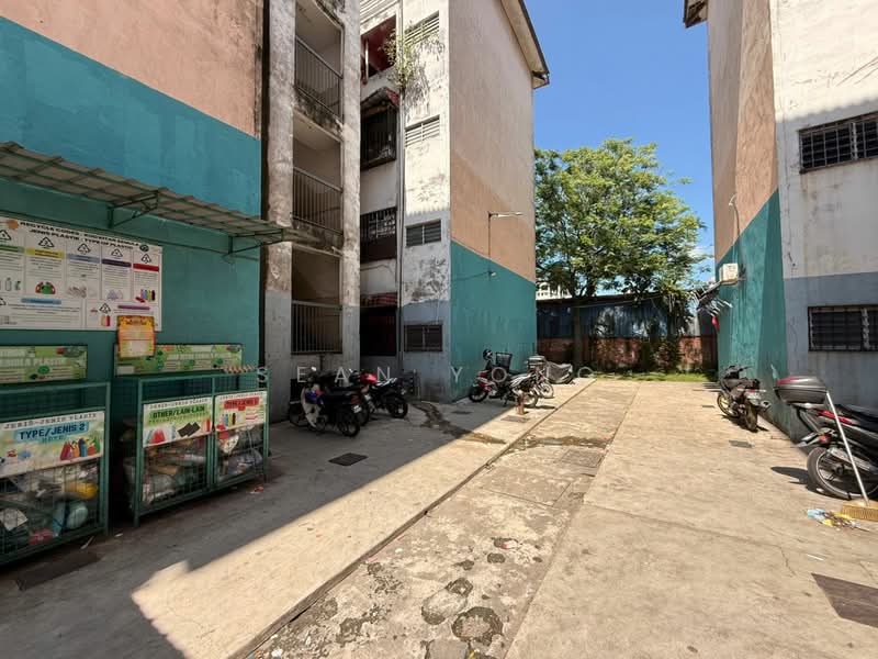 Exterior type: low-rise apartment block; features: two-storey building, exposed concrete facade, green and beige painted walls, balconies with metal railings, visible garbage disposal area, motorcycle parking area, paved walkway, surrounding greenery; style: minimalist.