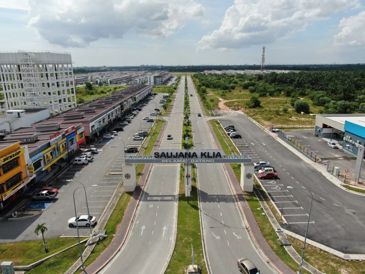 Exterior type: commercial development; features: wide asphalt road, multiple parking spaces, commercial shopfronts, signage archway, landscaped greenery, surrounding palm trees, adjacent industrial buildings, open space, utility poles, cloudy sky; style: modern.