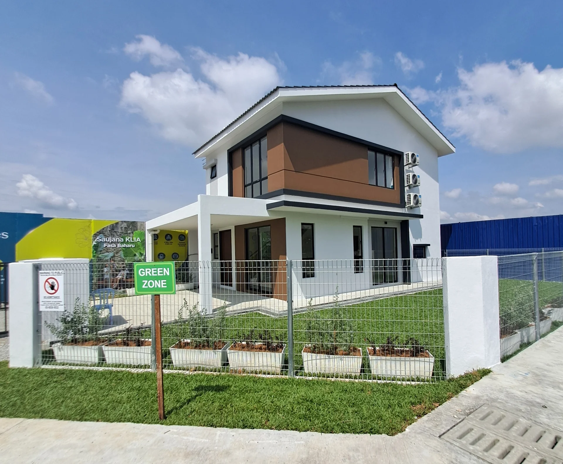 Exterior type: detached bungalow; features: white painted facade, brown accent panels, flat roof, approximately 2-storey, manicured lawn, fenced perimeter, multiple wall-mounted split AC units, landscaped garden beds, driveway with interlocking pavers, signage indicating 'GREEN ZONE', large windows, double-volume main entrance; style: modern.