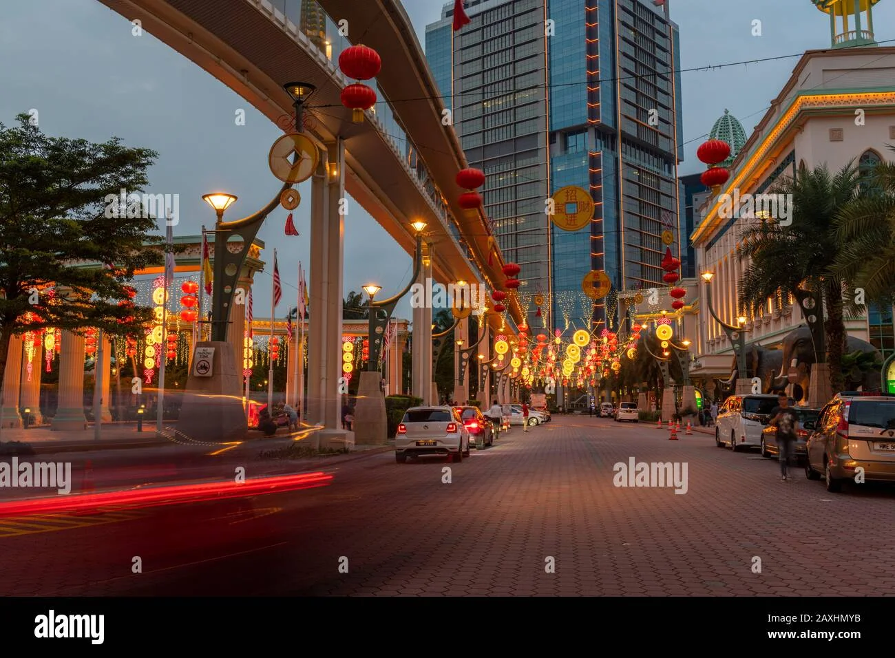 Exterior type: high-rise condominium; features: decorative lanterns, street lighting, paved walkway, surrounding palm trees, urban street view, multiple parked cars, overhead pedestrian bridge, festive decorations, modern glass facade, high-rise building with illuminated features; style: modern.