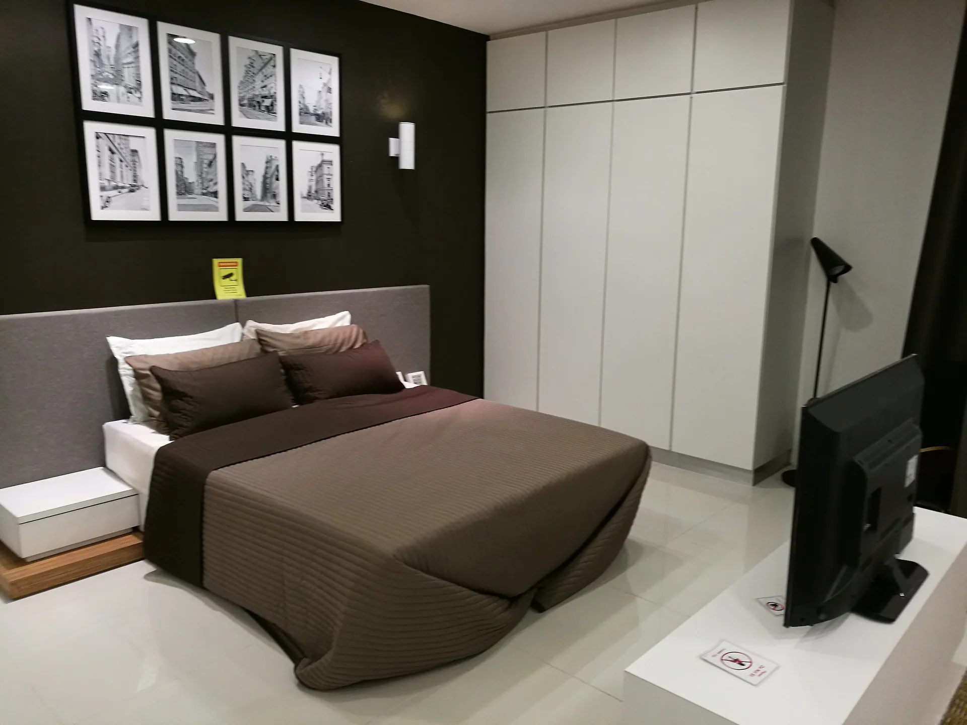 Bedroom features: queen-size bed with brown bedding, grey upholstered headboard, white bedside table, built-in wardrobe with white doors, wall-mounted artwork in black frames, recessed wall sconce, floor-to-ceiling mirror, TV on white console, laminate flooring, cream walls with dark accent wall, window with curtain treatment; style: modern.