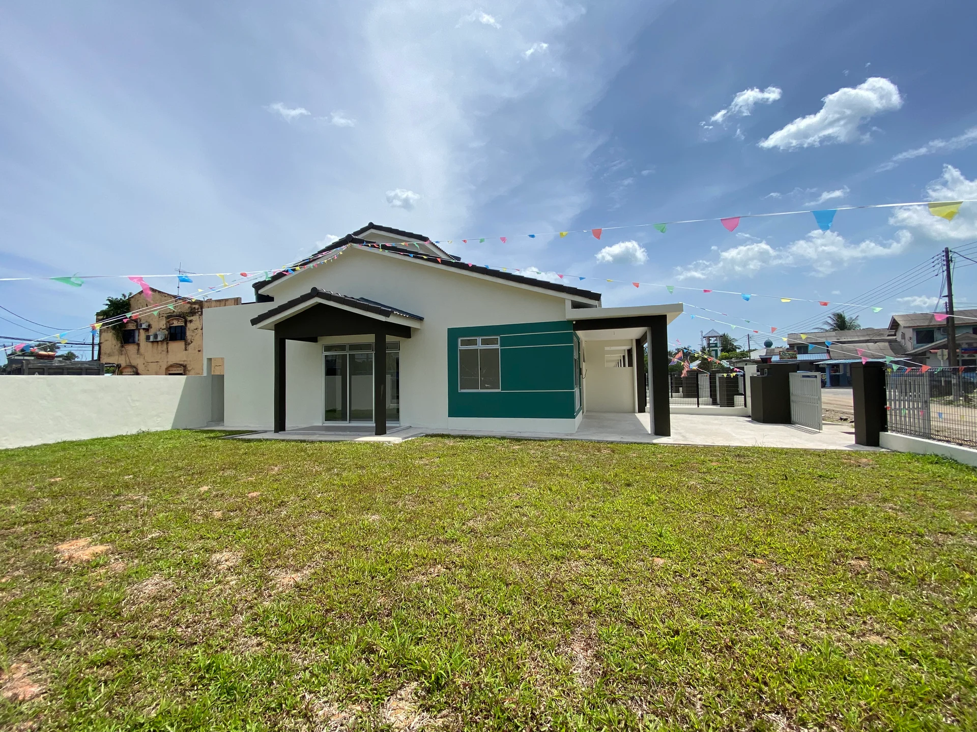 Exterior type: detached bungalow; features: flat roof, white painted facade, large windows, green accent wall, manicured lawn, paved driveway, decorative bunting, surrounded by residential area; style: modern.