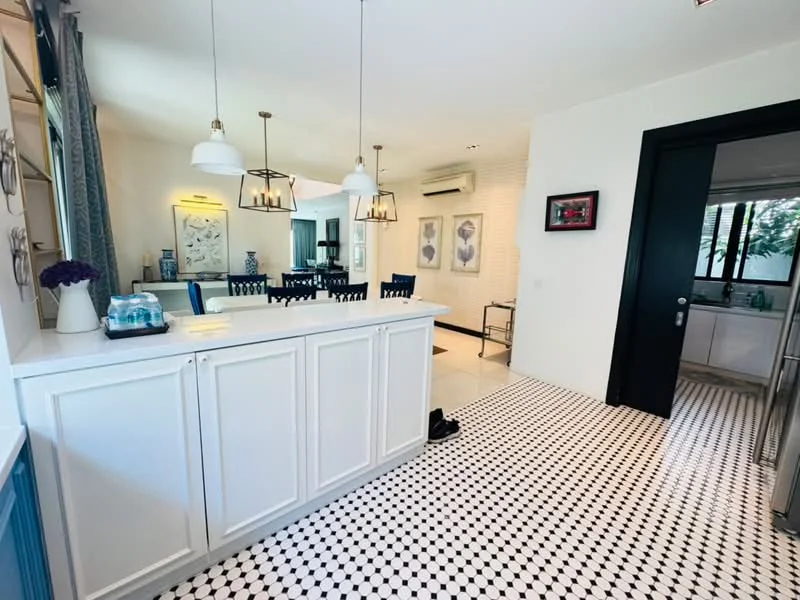 Kitchen features: white cabinetry, black and white checkered tile flooring, pendant lighting, kitchen island with seating, stainless-steel appliances, double-bowl stainless-steel sink, chrome pull-down faucet, open layout to dining area, wall-mounted split AC, decorative wall art; style: modern. Dining features: dining table with chairs, wall-mounted artwork, natural light from windows, air conditioning unit; style: modern. Room features: black door, window with view to outside.