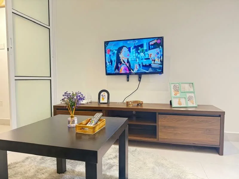 Living features: wall-mounted TV, wooden TV console, coffee table, decorative vase with flowers, remote control on table, framed artwork on table, light-colored walls, neutral tile flooring; style: modern.