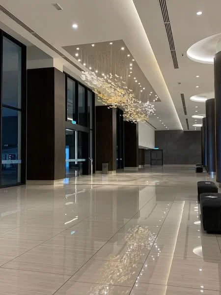 Room features: polished tile flooring, high ceiling with recessed lighting, chandelier with decorative elements, large floor-to-ceiling windows, dark wood paneling, modern wall sconces, open layout, neutral color palette; style: modern.