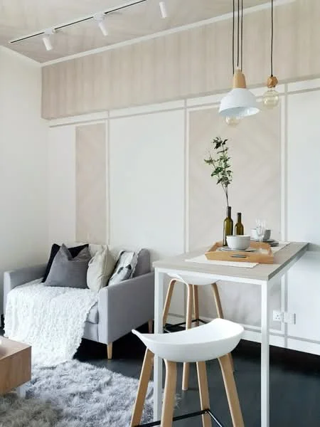 Living features: grey fabric sofa with cushions, white throw blanket, wooden coffee table, white bar table, white stool, hanging pendant light, track lighting, decorative vase with flowers, tableware on the bar table, textured wall panels, dark flooring, area rug; style: modern.