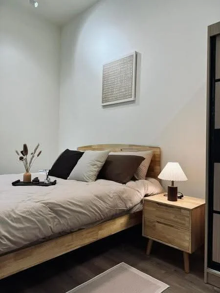 Bedroom features: queen-size bed with beige bedding, wooden bed frame, two decorative pillows in brown and cream, nightstand with wooden finish, table lamp with white shade, framed artwork on wall, wooden flooring, area rug with light color; style: modern.