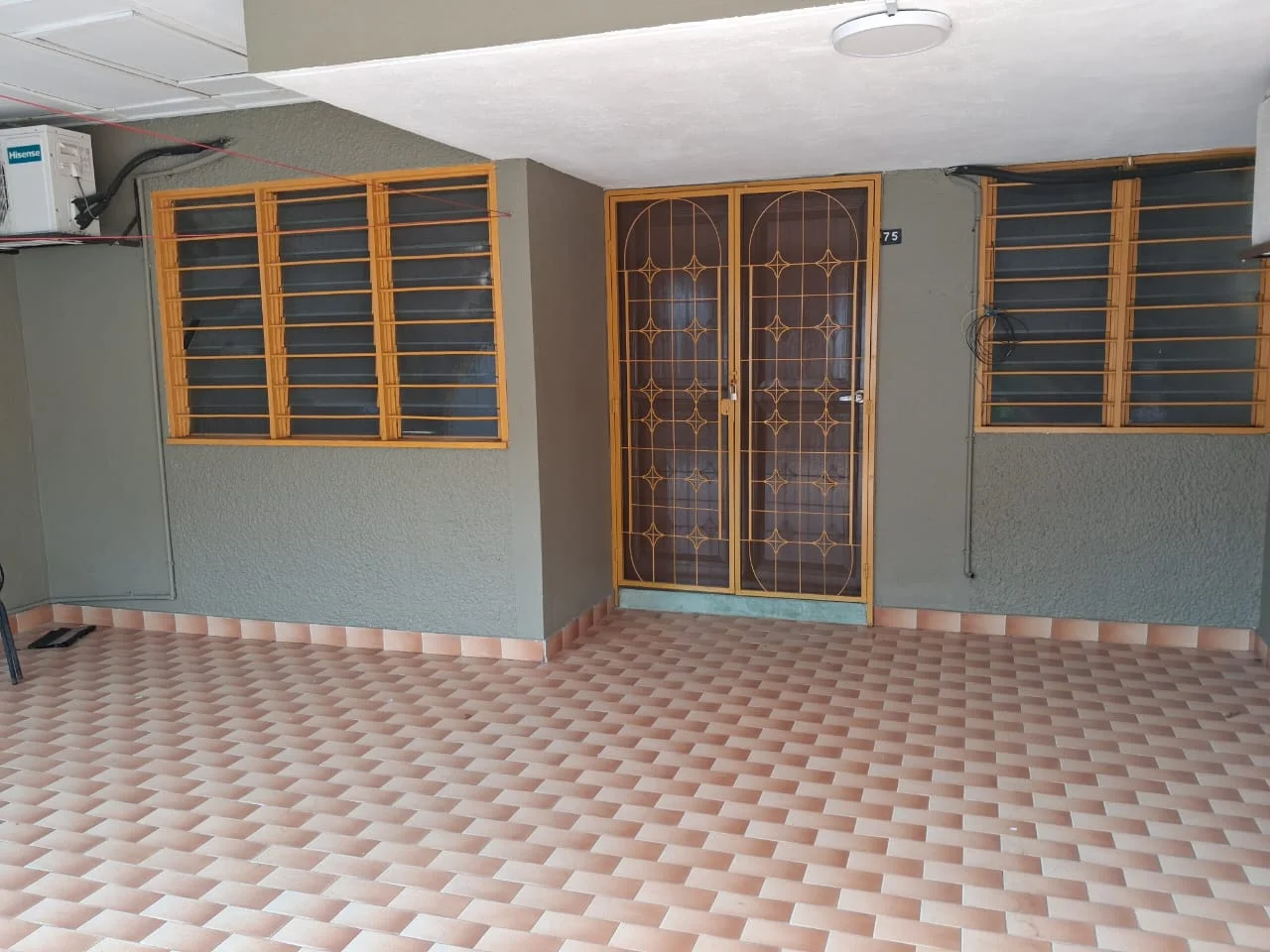 Exterior features: double-door entrance with decorative grille, grey painted walls, tan and cream patterned floor tiles, two windows with metal grilles, wall-mounted air conditioning unit, overhead ceiling light, exposed electrical wiring, covered porch area.