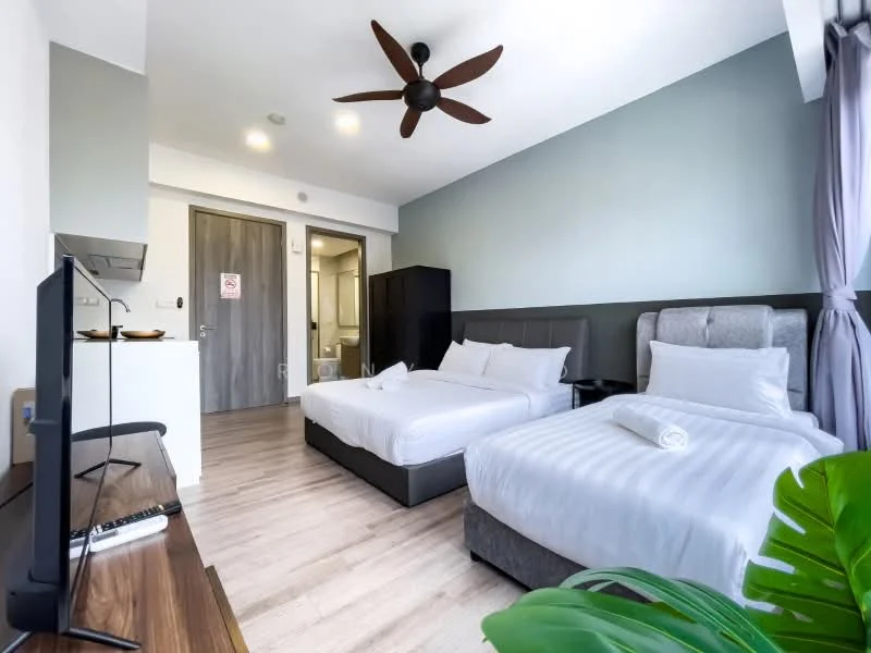 Bedroom features: two single beds with white bedding, upholstered headboard, wooden flooring, 5-blade ceiling fan, wall-mounted TV, desk with chair, large window with sheer curtains, decorative plant, built-in wardrobe, light grey accent wall, recessed ceiling lights; style: modern. Kitchenette features: compact kitchen counter, stainless-steel sink, built-in cabinets, microwave, mini fridge; style: modern. Bathroom features: shower area, toilet, vanity with mirror, tile flooring, white walls; style: modern.
