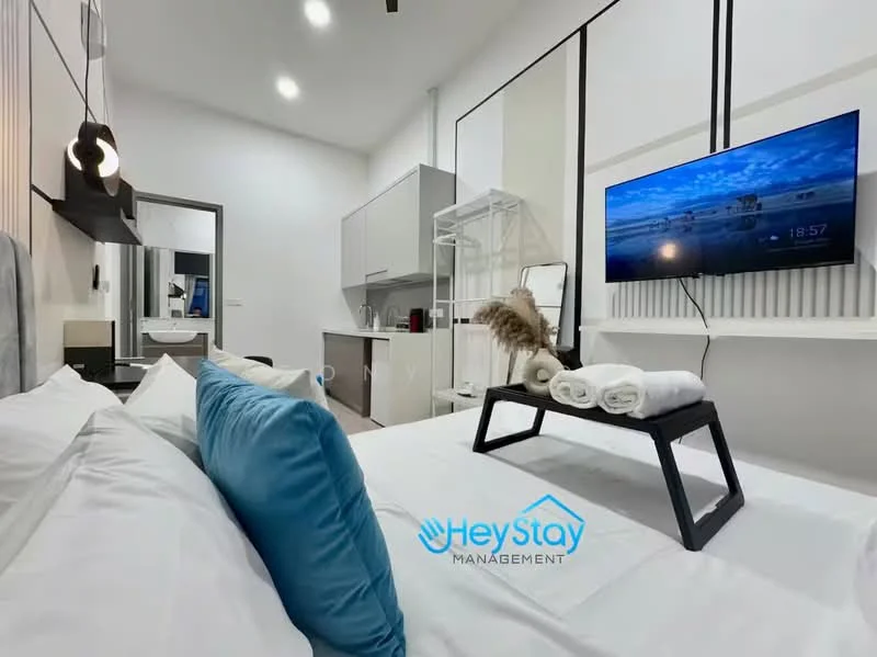 Bedroom features: queen-size bed with white bedding, blue decorative pillow, wall-mounted TV, black coffee table, white towels on coffee table, grey accent wall, white painted walls, recessed lighting, ceiling fan, open shelving unit, kitchenette area with grey cabinets, mirror on wall, wooden flooring; style: modern.
