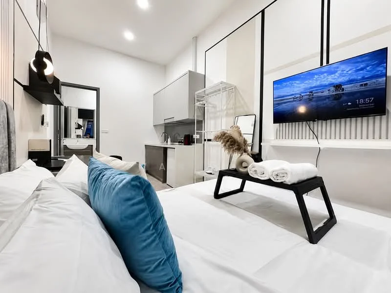 Room features: queen-size bed with white bedding, blue decorative pillow, wall-mounted TV, black TV console, open shelving unit, kitchenette with grey cabinetry, sink, refrigerator, white walls, recessed lighting, wooden flooring, decorative plant, towels on a black tray; style: modern.