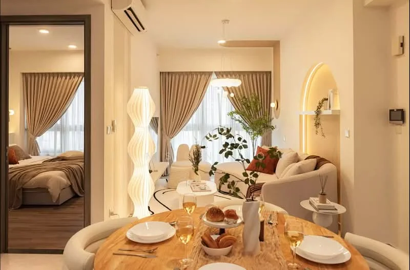 Dining features: round wooden dining table, 4 upholstered dining chairs, table set with wine glasses and plates, decorative plant in vase, croissants on plate, soft lighting from pendant lamp; style: modern; view: living. Living features: L-shape sectional sofa in cream, decorative throw pillows, coffee table, floor lamp, large windows with sheer curtains, decorative plants, wall-mounted shelves with decor, soft lighting, wooden flooring; style: modern. Bedroom features: king-size bed with beige bedding, upholstered headboard, bedside lamps, large window with curtains, decorative plants, soft lighting; style: modern.