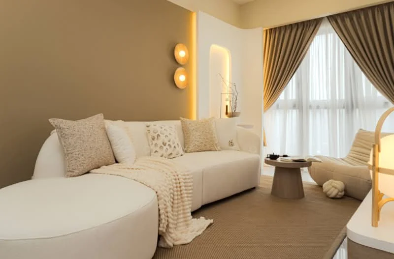 Living features: curved white fabric sofa, beige throw blanket, textured white cushions, accent wall in taupe, recessed lighting, two wall sconces, large floor-to-ceiling windows with sheer curtains, round coffee table, beige area rug, decorative plant, soft lighting, neutral color palette; style: modern.