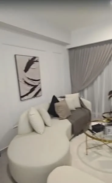 Living features: curved white sectional sofa, decorative throw pillows in black and beige, round coffee table with gold frame, framed abstract artwork on wall, sheer curtains, light-colored carpet, wall-mounted lighting, white painted walls; style: modern.