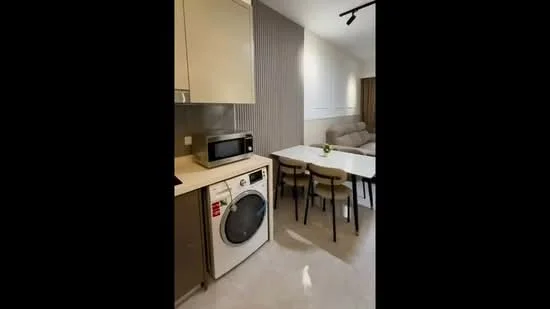 Room features: white walls, light grey accent wall, laminate flooring, kitchen cabinetry in light colour, microwave, front-load washing machine, dining table with four chairs, sofa in neutral colour, recessed downlights, open layout; style: modern.