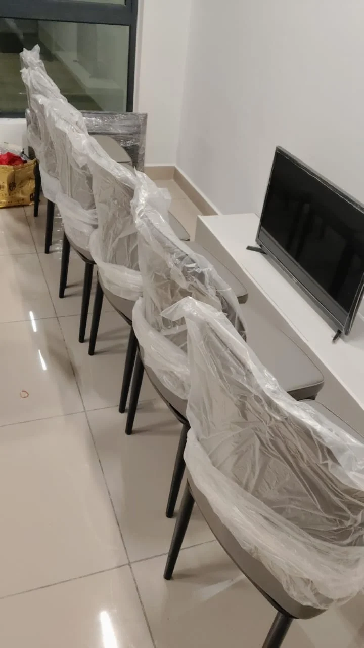 Room features: plastic-wrapped dining chairs, flat-screen TV, light-colored tiled flooring, white painted walls, minimalist decor, small console table, large window, neutral color palette; style: modern.