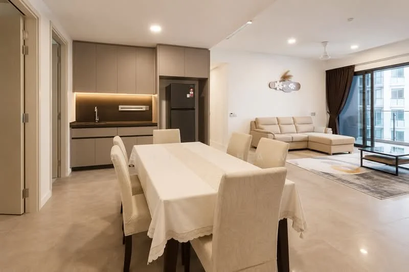 Dining features: 6-seater dining table with white tablecloth, upholstered dining chairs in cream, pendant light above dining table, floor-to-ceiling windows, grey tiled flooring, neutral painted walls, recessed downlights, open layout to kitchen and living area; style: modern; view: city. Kitchen features: kitchen cabinets in grey, black countertop, stainless-steel fridge, built-in oven, single-bowl stainless-steel sink, chrome pull-down faucet, ceramic tile backsplash, recessed downlights; style: modern. Living features: L-shape sectional sofa in beige, coffee table, area rug with circular pattern, TV console, wall-mounted TV, floor-to-ceiling windows, curtains in dark brown, neutral painted walls; style: modern; view: city.