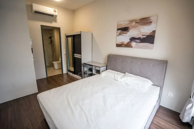 Bedroom features: queen-size bed with white bedding, upholstered headboard in grey, wooden flooring, wall-mounted air conditioning unit, framed artwork on wall, built-in wardrobe with sliding doors, study desk, chair, en-suite bathroom visible; style: modern. Bathroom features: toilet, shower with glass enclosure, tile flooring, wall-mounted shower head.
