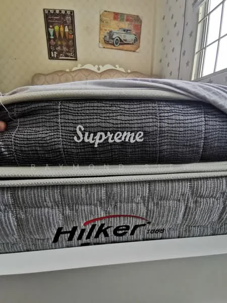 Room features: mattress with 'Supreme' branding, mattress with 'Hiker 1699' branding, bed frame, decorative wall with patterned wallpaper, window with natural light, wall art or poster, light-colored bedding.