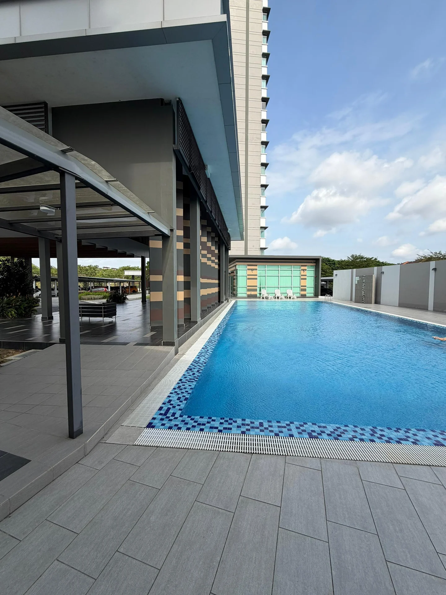 Exterior type: high-rise condominium; features: infinity pool, pool deck with loungers, glass curtain wall, covered outdoor dining area, manicured landscaping, tiled flooring around pool, modern architectural design, surrounded by greenery; style: modern.