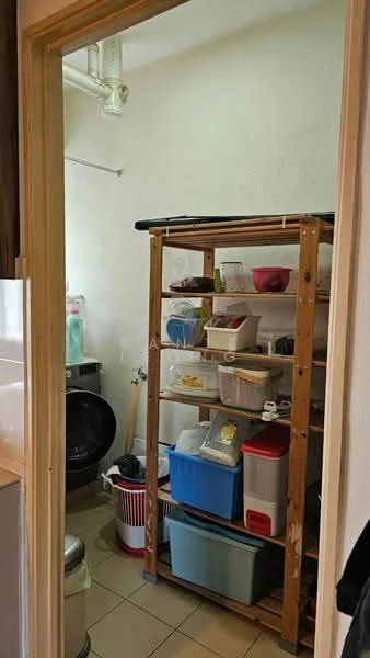 Laundry features: wooden shelving unit, plastic storage containers, metal trash bin, washing machine, tiled flooring, white painted walls, light fixture on ceiling.