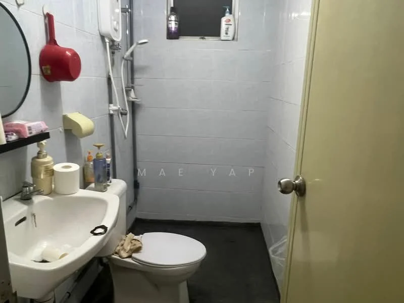 Bathroom features: white ceramic toilet, single-sink vanity with wall-mounted mirror, chrome basin mixer faucet, shower with handheld showerhead, white tiled walls, dark tiled flooring, soap dispenser, toilet paper holder, window for natural light, bottle of shampoo, bottle of body wash, tissue box.