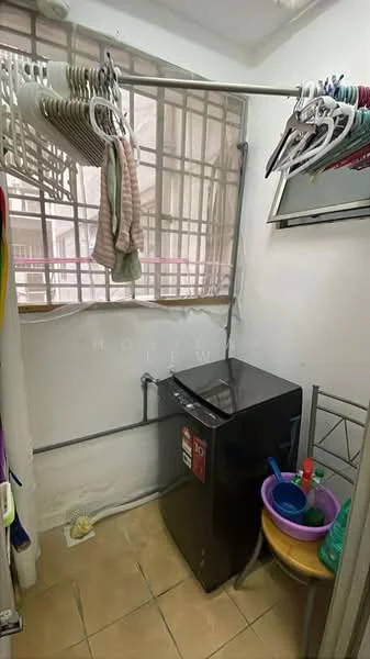 Laundry features: front-load washing machine in black, clothes drying rack, tile flooring, window with metal grille, plastic storage container, cleaning supplies, hanging clothes, shelf with metal frame.