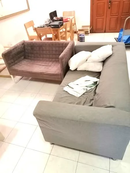 Living features: 3-seater fabric sofa in grey, 2-seater fabric sofa in brown, white throw pillows, folding dining table with wooden chairs, tile flooring, neutral wall color, stack of magazines or papers on the sofa; style: modern.