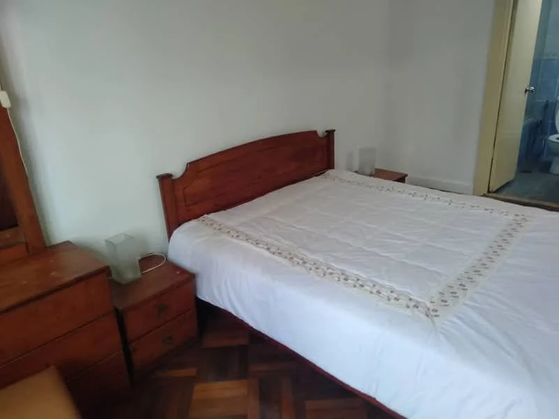 Bedroom features: queen-size bed with white bedding, wooden headboard, two bedside tables, lamps on bedside tables, parquet wood flooring, white painted walls, wardrobe visible in background, doorway leading to bathroom; style: traditional.