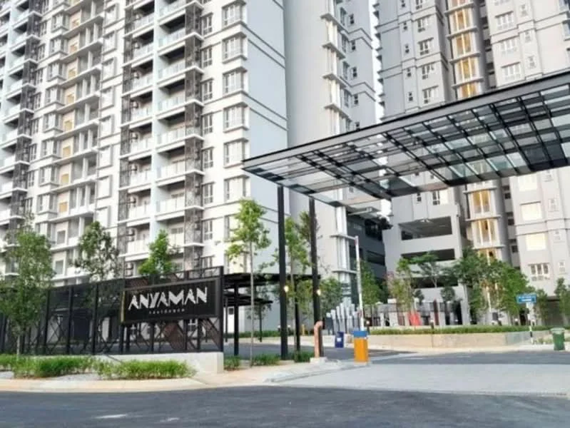 Exterior type: high-rise condominium; features: glass canopy entrance, gated entry, landscaped garden, manicured lawn, modern facade with grey and white colors, visible parking area, security guard house, signage with 'ANYAMAN' name, surrounded by greenery; style: modern.