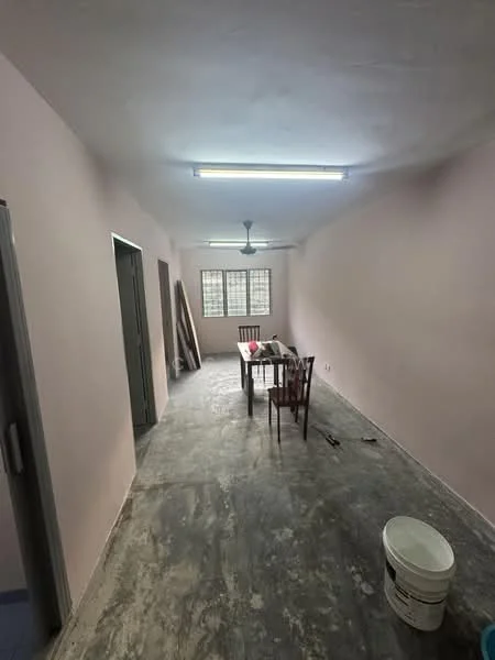Room features: pink painted walls, concrete flooring, ceiling with fluorescent lighting, ceiling fan, wooden dining table, four wooden chairs, window with grid pattern, bucket on the floor, unfinished space with construction materials.