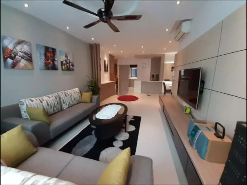Living features: 3-seater fabric sofa in grey, 2-seater fabric sofa in grey, yellow decorative cushions, round coffee table with dark wood finish, area rug with geometric pattern in black and white, wall-mounted TV, entertainment unit with light wood finish, decorative wall art with colorful designs, ceiling fan with light, recessed downlights, plant in ceramic pot, open shelving unit, clock on wall, kitchen area visible in background; style: modern.