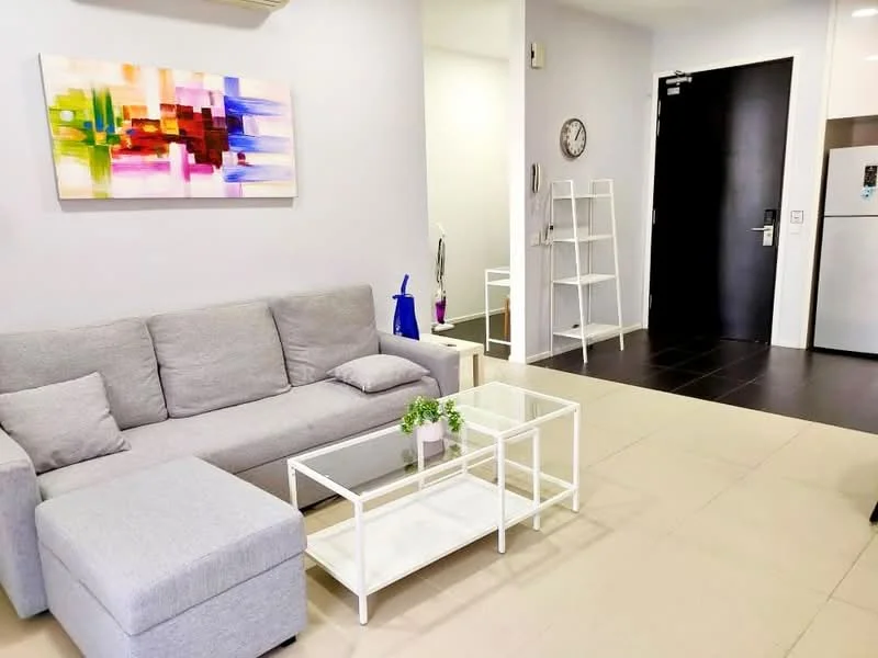 Living features: grey fabric sofa, white coffee table with glass top, decorative plant on coffee table, wall-mounted clock, abstract artwork on wall, open shelving unit, floor tiles in light beige, dark wood flooring near entrance, wall-mounted air conditioning unit, entrance door, visible kitchen area in background; style: modern.