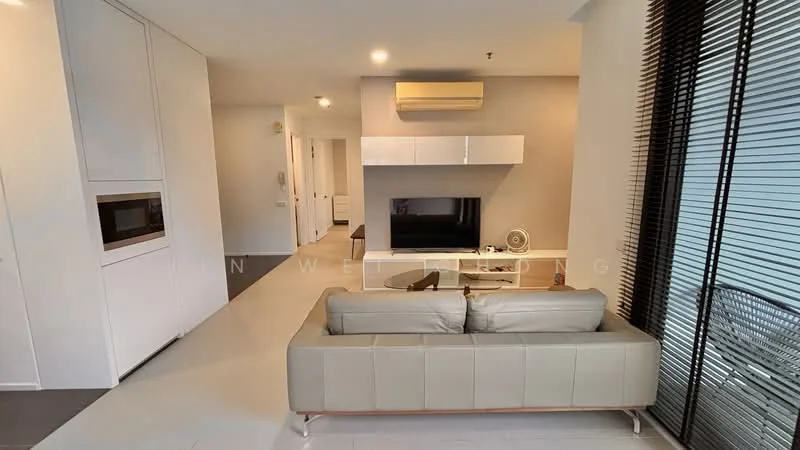 Living features: 3-seater leather sofa in light beige, wall-mounted TV, white TV console, built-in cabinetry, air conditioning unit, large window with sheer curtains, light-colored flooring, decorative wall paneling, ceiling with recessed lighting; style: modern. Room features: white painted walls, open layout, kitchenette area with built-in microwave.