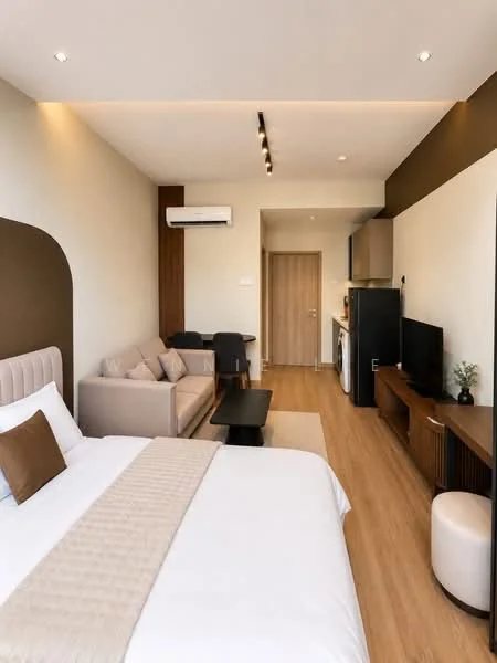 Room features: queen-size bed with beige headboard, white bedding with decorative throw, wooden flooring, sofa in light grey, black coffee table, wall-mounted TV, dark wood TV console, refrigerator, washing machine, air conditioning unit, recessed ceiling lights, neutral wall colors, decorative plant on console; style: modern.