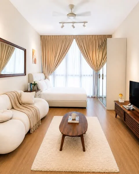 Bedroom features: queen-size bed with upholstered headboard, 2-seater fabric sofa in white, wooden coffee table, area rug with neutral color, floor-to-ceiling windows with sheer curtains, light brown painted walls, wooden flooring, ceiling fan with integrated light, wall-mounted TV, framed mirror, decorative plant on side table; style: modern.