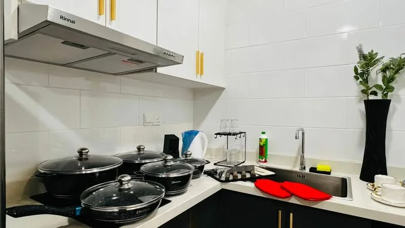 Kitchen features: stainless-steel sink, chrome pull-down faucet, white tiled backsplash, white cabinets with gold handles, black countertop, black cooking pots with lids, glass kettle, dish drying rack, red oven mitts, decorative plant in black vase, built-in range hood; style: modern.