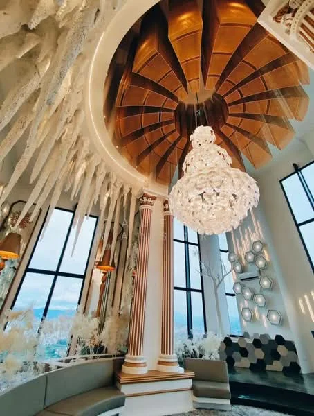 Room features: chandelier with crystal design, exposed wooden ceiling with geometric pattern, large floor-to-ceiling windows, decorative columns, modern wall decor with hexagonal shapes, curved seating area, white decorative elements, ambient lighting; style: modern.