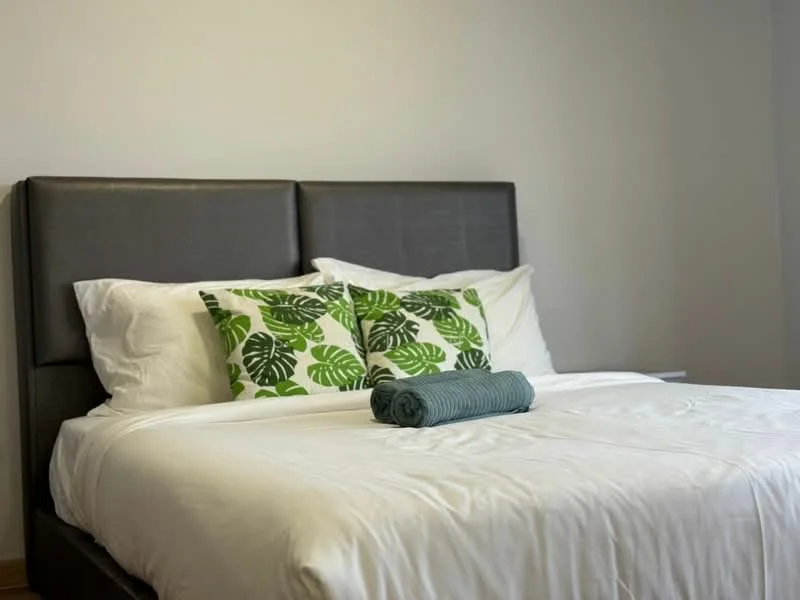 Bedroom features: king-size bed, upholstered headboard in dark grey, white bedding, green leaf-patterned decorative pillows, rolled towel on bed, light grey walls, wooden flooring; style: modern.