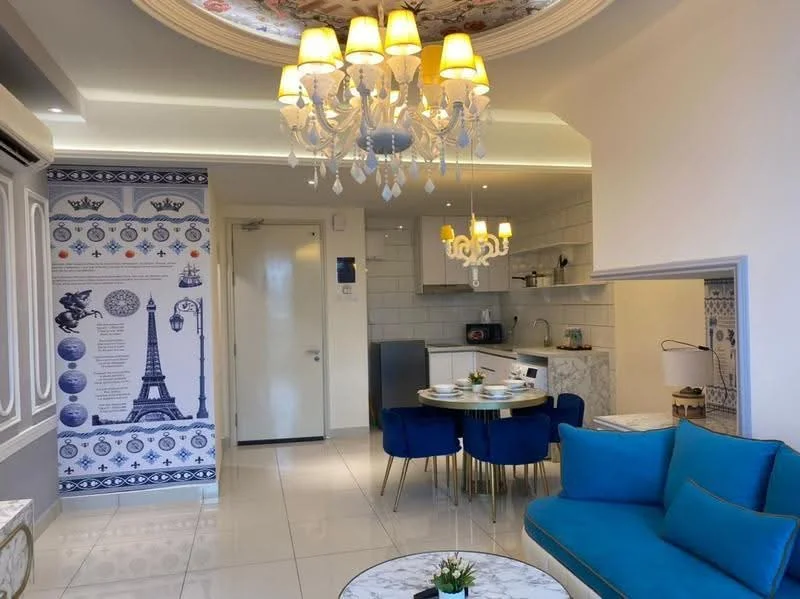 Living features: white painted walls, glossy tile flooring, chandelier with multiple bulbs, 5-blade ceiling fan with light, blue fabric sofa, round marble coffee table, decorative wall with Eiffel Tower design, dining table with four chairs, built-in kitchen with white cabinetry, microwave, stainless-steel sink, kitchen countertop, recessed lighting; style: modern.