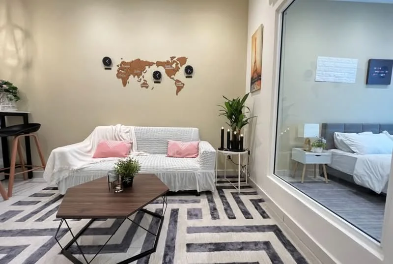 Living features: white fabric sofa with throw blanket, pink decorative pillows, wooden coffee table, geometric patterned area rug, wall-mounted world map decor, potted plant, floor-to-ceiling window, grey upholstered bed visible through window, white bedding on bed, decorative wall art; style: modern.