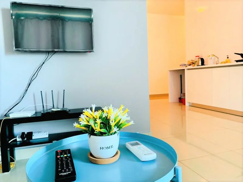 Living features: flat-screen TV mounted on wall, black TV console, remote control, decorative plant in pot, round coffee table in blue, white walls, tile flooring, open kitchen view in background, kitchen appliances visible, kitchen cabinets, kitchen countertop; style: modern.