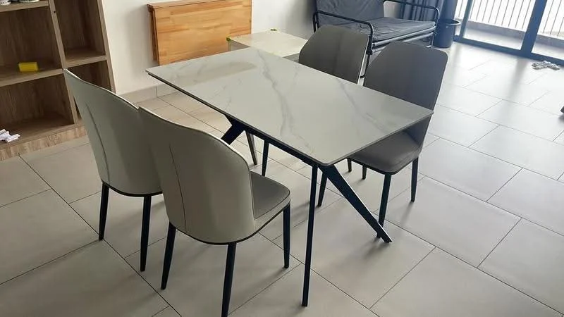 Dining features: rectangular dining table with marble top, 4 dining chairs with grey upholstery, metal legs on table and chairs, light-coloured tiled flooring, wooden sideboard in background, large windows with natural light, minimalist decor; style: modern.