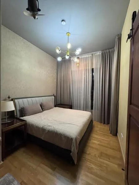 Bedroom features: queen-size bed, upholstered headboard, wooden bedside table, table lamp, curtains, wooden flooring, chandelier with multiple bulbs, wall-mounted light fixture, decorative wall, floor-to-ceiling window; style: modern.