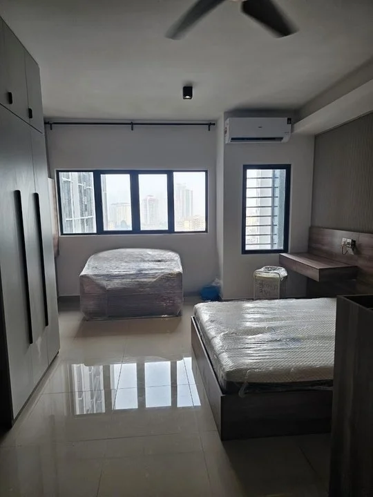 Bedroom features: two beds with mattresses wrapped in plastic, built-in wardrobe with sliding doors, study desk, wall-mounted air conditioning unit, floor-to-ceiling windows, white painted walls, light-colored tile flooring, ceiling fan with light, window with horizontal slats; style: modern; view: city.