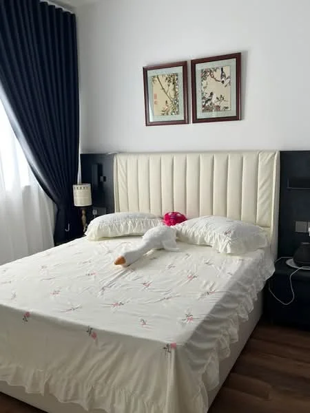 Bedroom features: king-size bed with upholstered headboard, white bedspread with floral embroidery, two bedside tables, framed artwork on walls, floor-to-ceiling curtains in dark blue, laminate wood flooring, night lamp on bedside table, decorative plush toy on bed; style: modern.