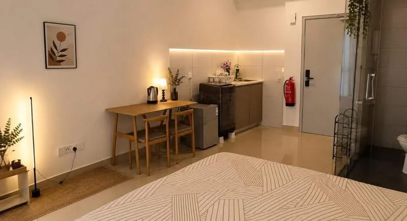 Room features: queen-size bed, wooden flooring, white painted walls, recessed LED downlights, table with two chairs, small fridge, kettle, decorative artwork, fire extinguisher, glass shower enclosure, sink with countertop, wall-mounted mirror, plant decor, area rug; style: modern.