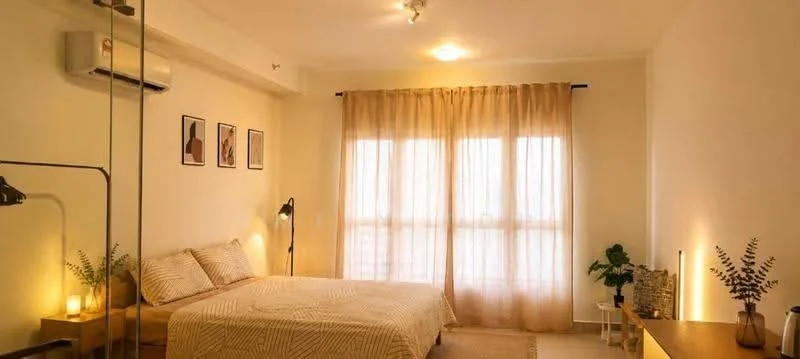 Bedroom features: queen-size bed, upholstered headboard, 2 bedside lamps, floor-to-ceiling windows, sheer curtains, 1 wall-mounted split AC, wooden flooring, decorative plants in ceramic pots, framed artwork on walls, light-colored bedding, wooden side table, recessed ceiling lights; style: modern.
